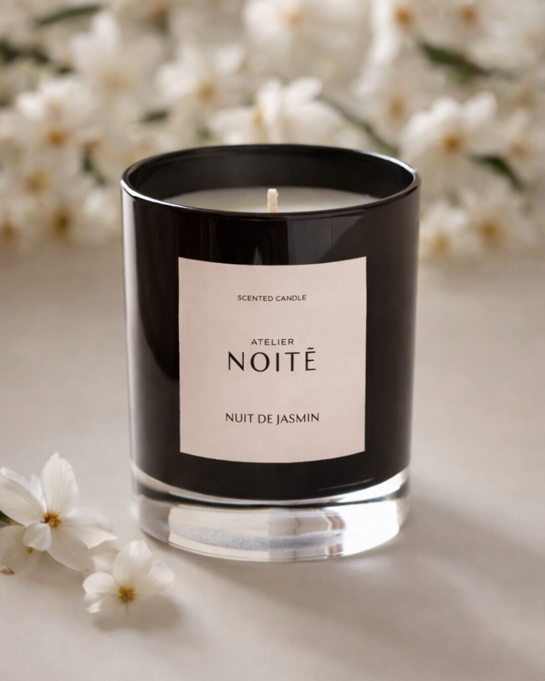 Scented candle with 'Atelier Noitē' label on a light surface with white jasmine flowers in the background