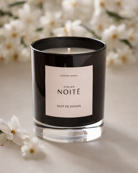Scented candle with 'Atelier Noitē' label on a light surface with white jasmine flowers in the background