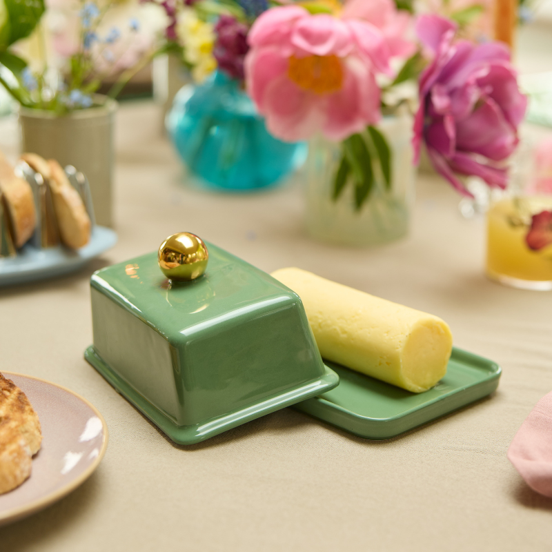 Green butter dish with a gold knob on the lid that is off the tray to reveal a yellow stick of butter inside on a table with pastries and flowers in the background.