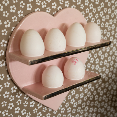Pink heart-shaped egg holder with eggs hung on a beige floral patterned wall.