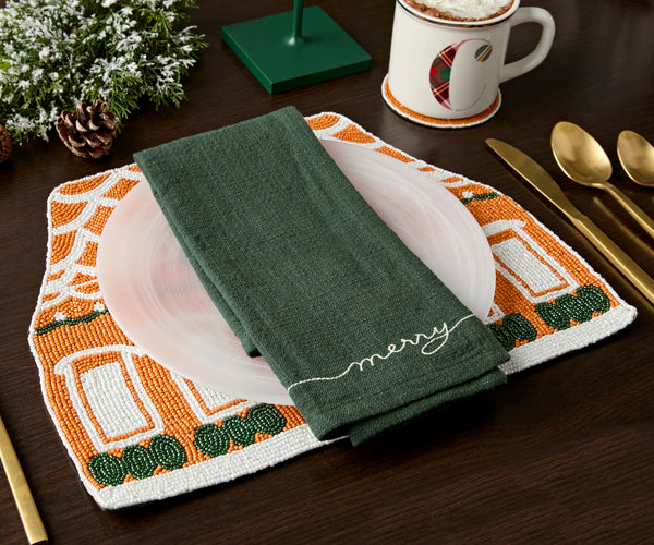 Gingerbread house beaded placemat under a white plate and green napkin, with a holiday mug on a beaded coaster