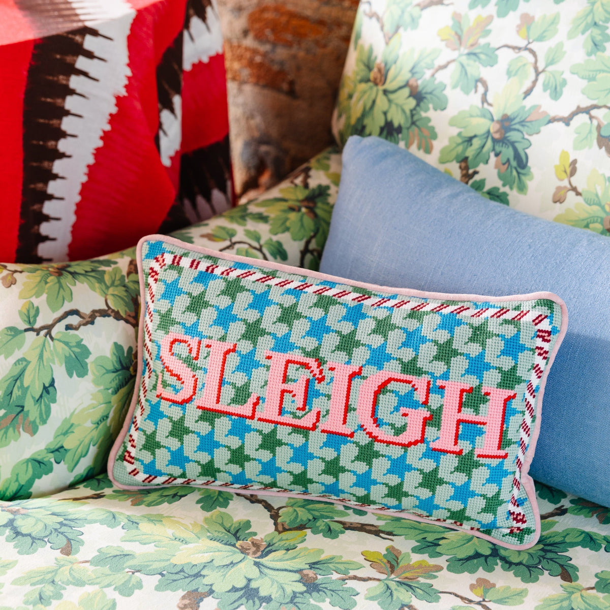 Decorative pillow with 'SLEIGH' text on a floral-patterned couch against a brick wall.