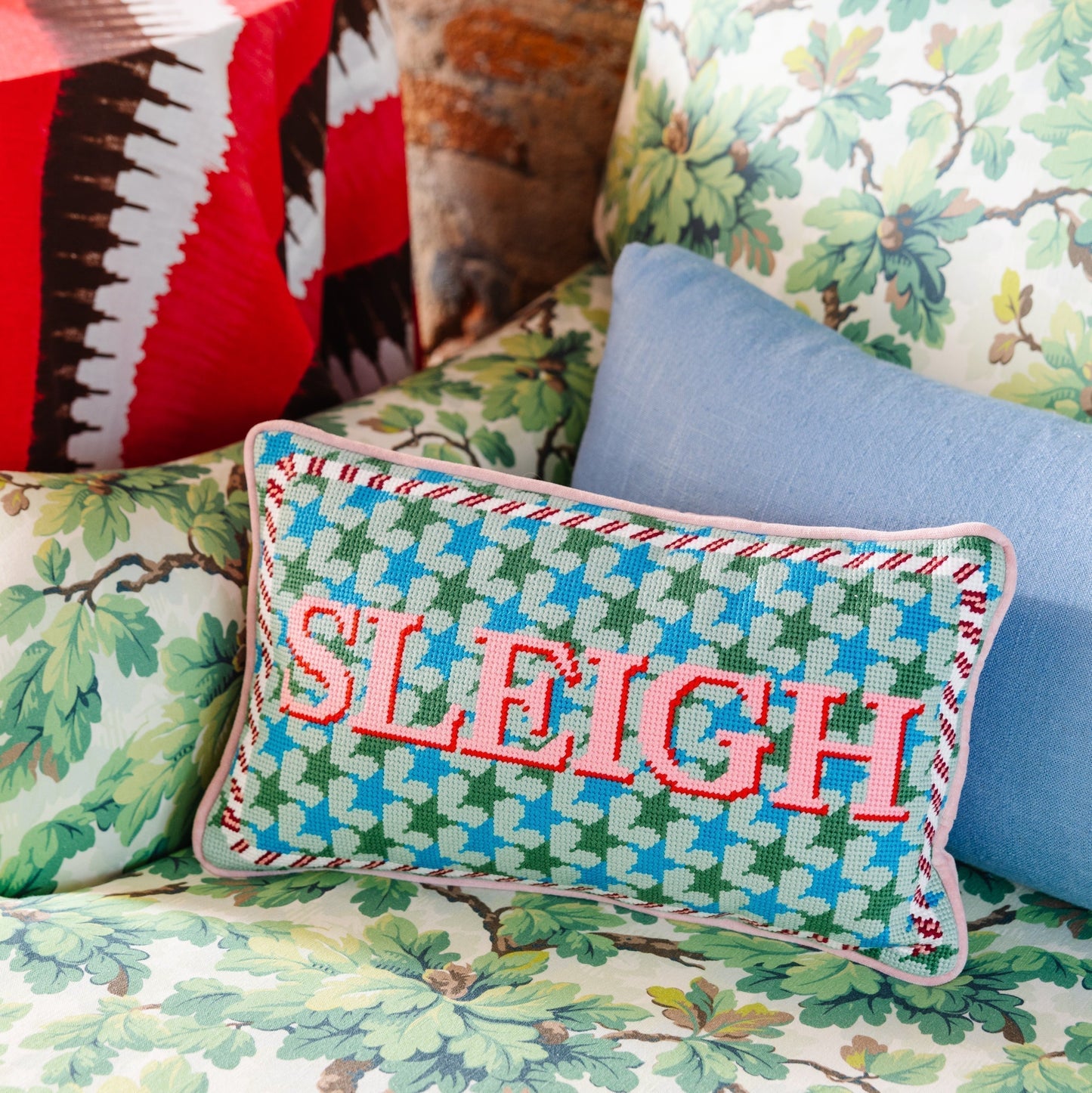 Decorative pillow with 'SLEIGH' text on a floral-patterned couch against a brick wall.
