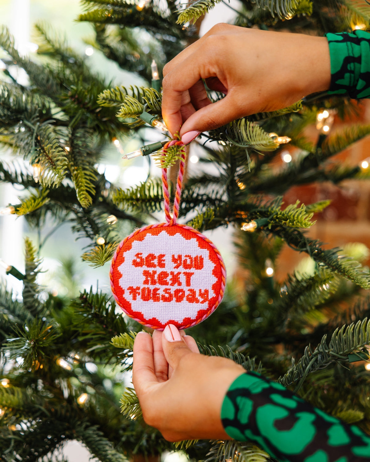 Person hanging a red ornament with 'See you next Tuesday' text on a Christmas tree.