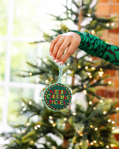 Hand holding a 'Merry Christmas Bitch' ornament in front of a decorated Christmas tree.