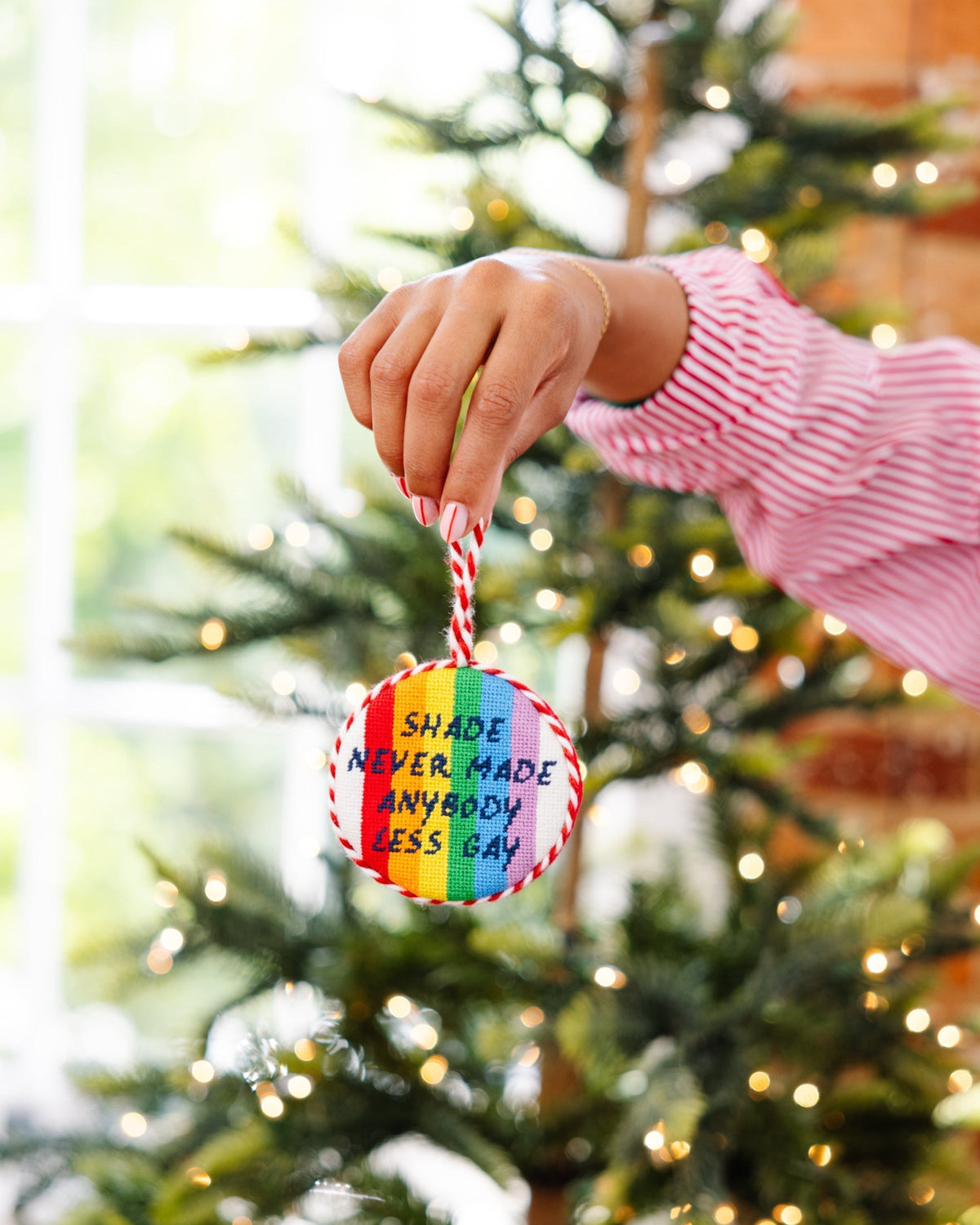 Hand holding a rainbow-colored ornament with text in front of a decorated Christmas tree.