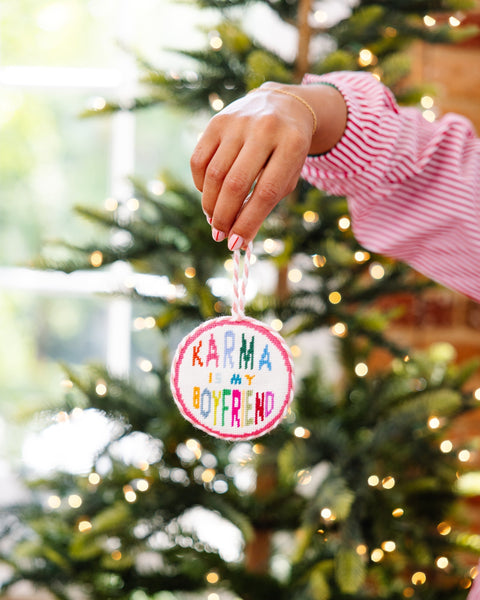 Hand holding a Christmas ornament with 'Karma is my boyfriend' text in front of a decorated tree.