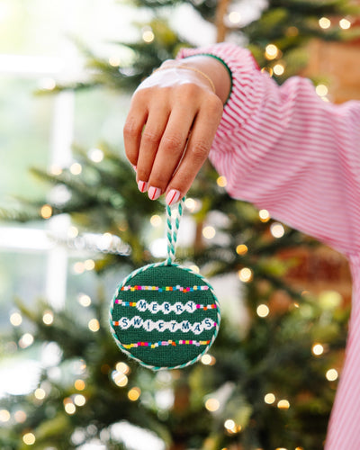 Hand holding a Christmas ornament with 'Meowy Christmas' text in front of a decorated tree.