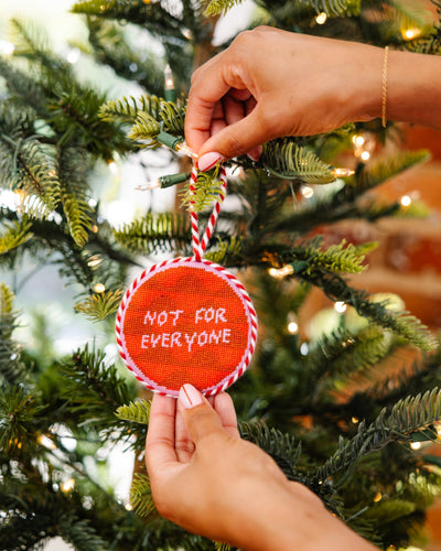 Hand holding a red ornament with 'Not for Everyone' text on a decorated Christmas tree.