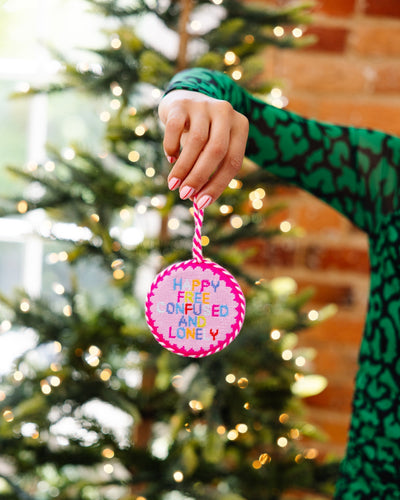 Hand holding a colorful ornament with text in front of a Christmas tree