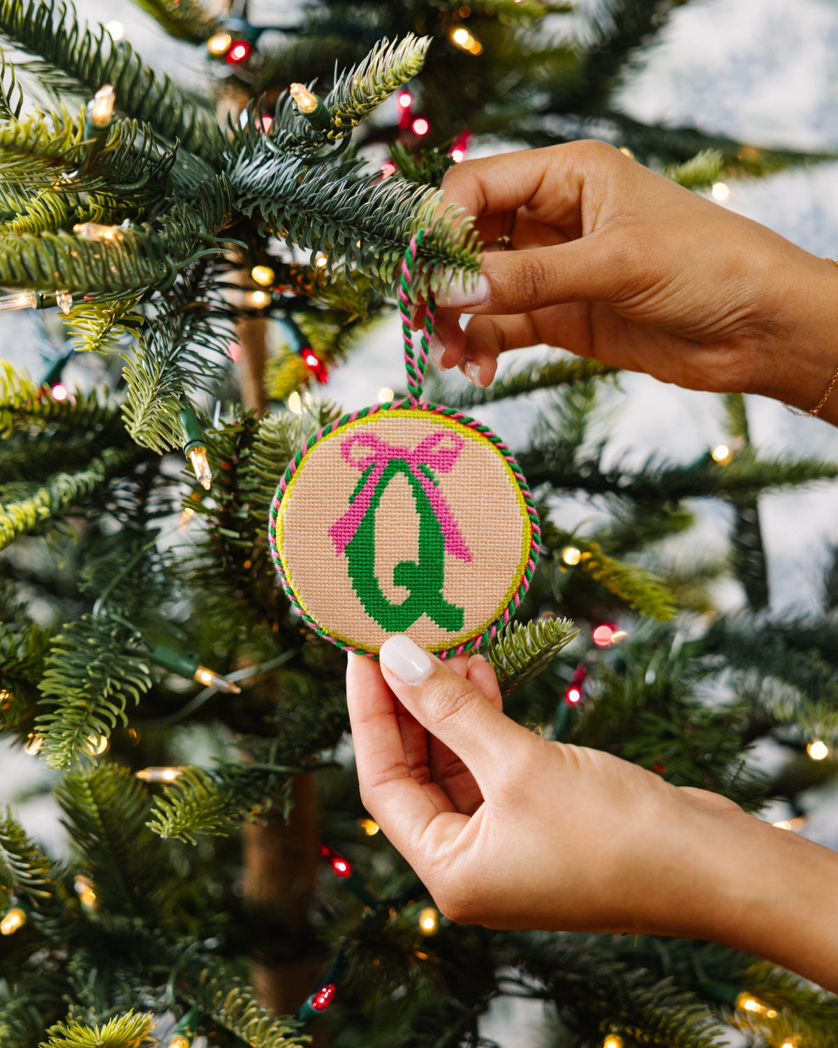 Hand holding a Christmas ornament with a letter 'Q' and pink bow on a decorated tree.