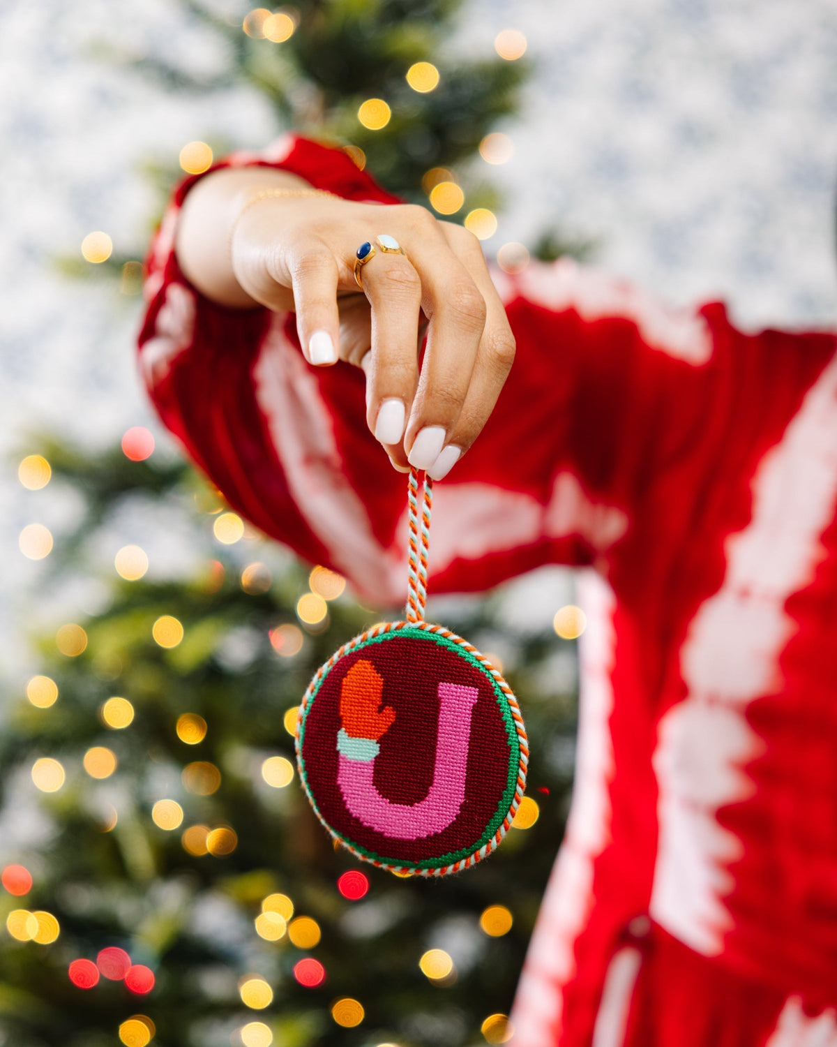 Person wearing a red sweater holding a Christmas ornament with a letter 'J' in front of a decorated tree.