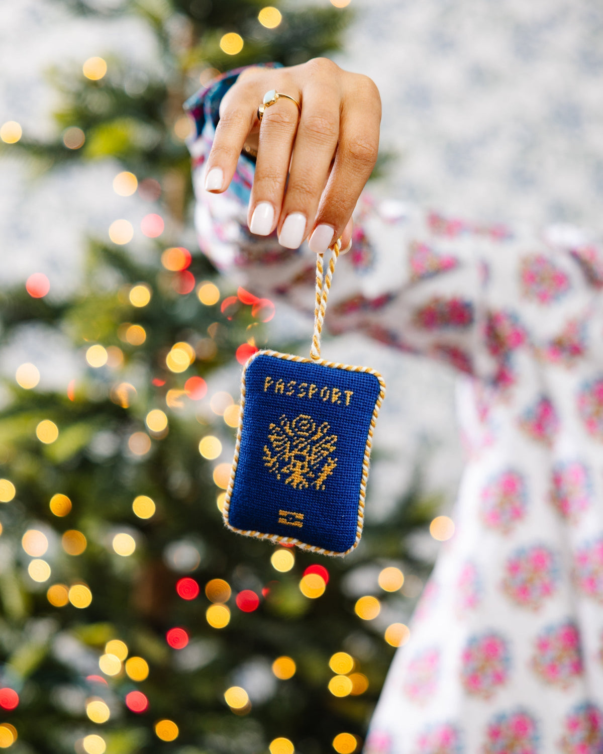 Hand holding a blue passport-shaped keychain in front of a blurred Christmas tree with lights.