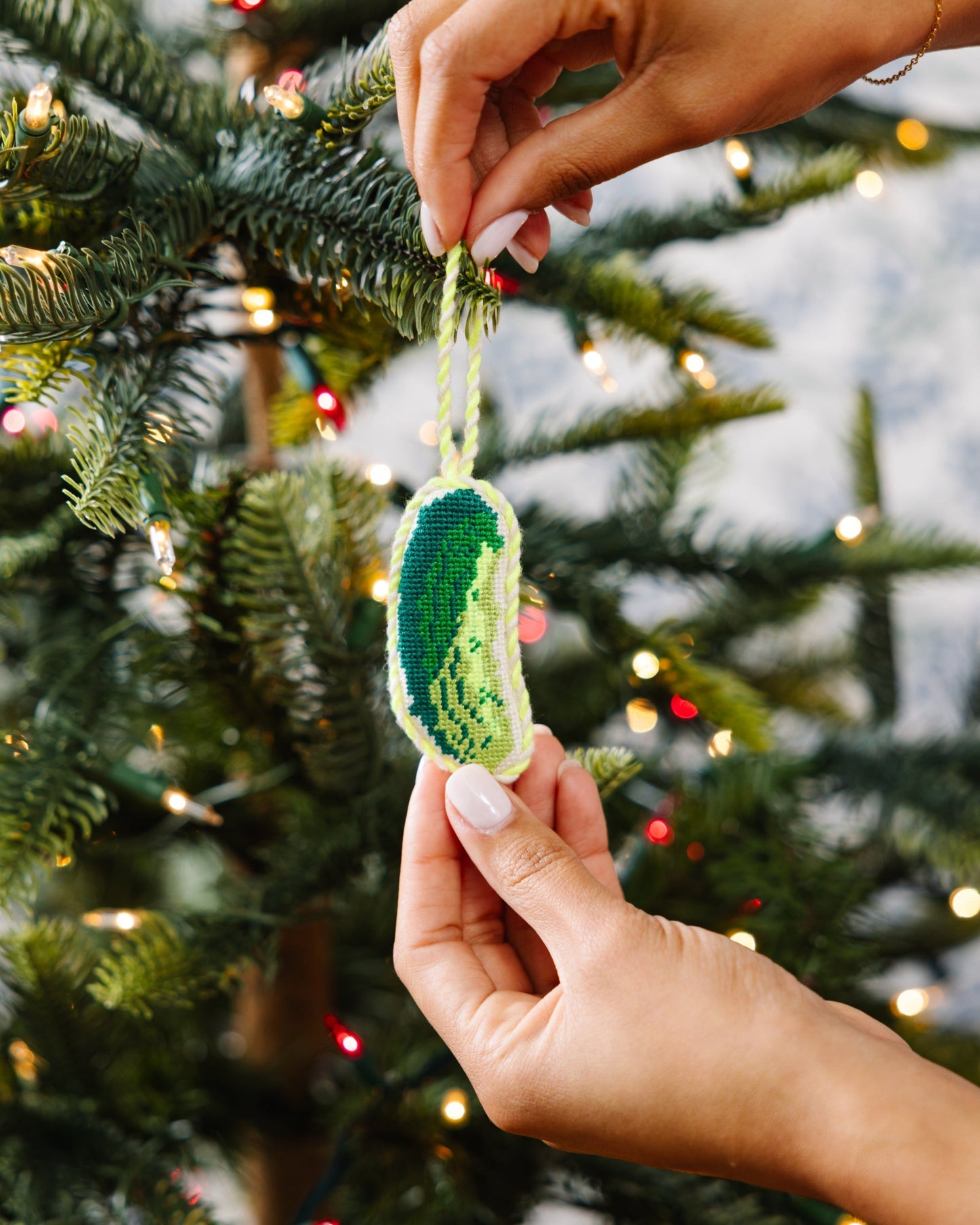Hand holding a green and blue knitted ornament in front of a decorated Christmas tree.