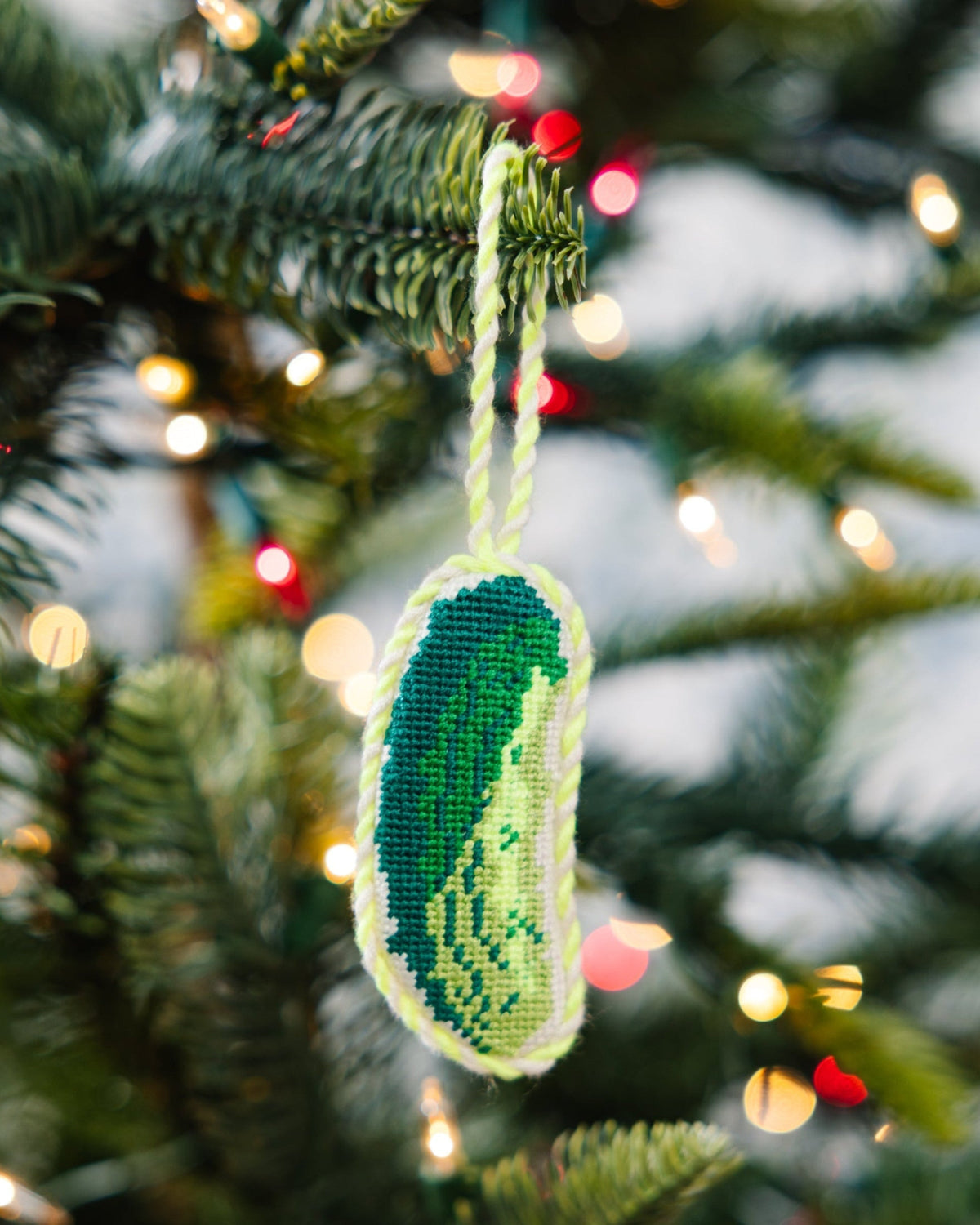 Decorative ornament shaped like a pickle hanging on a Christmas tree with lights in the background.