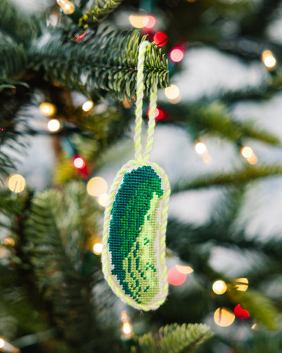Decorative ornament shaped like a pickle hanging on a Christmas tree with lights in the background.