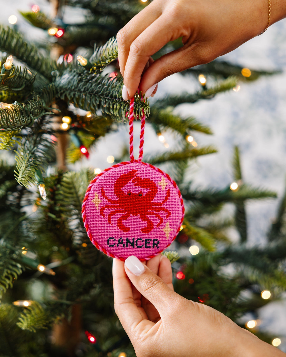Hand holding a pink 'Cancer' zodiac ornament against a Christmas tree background
