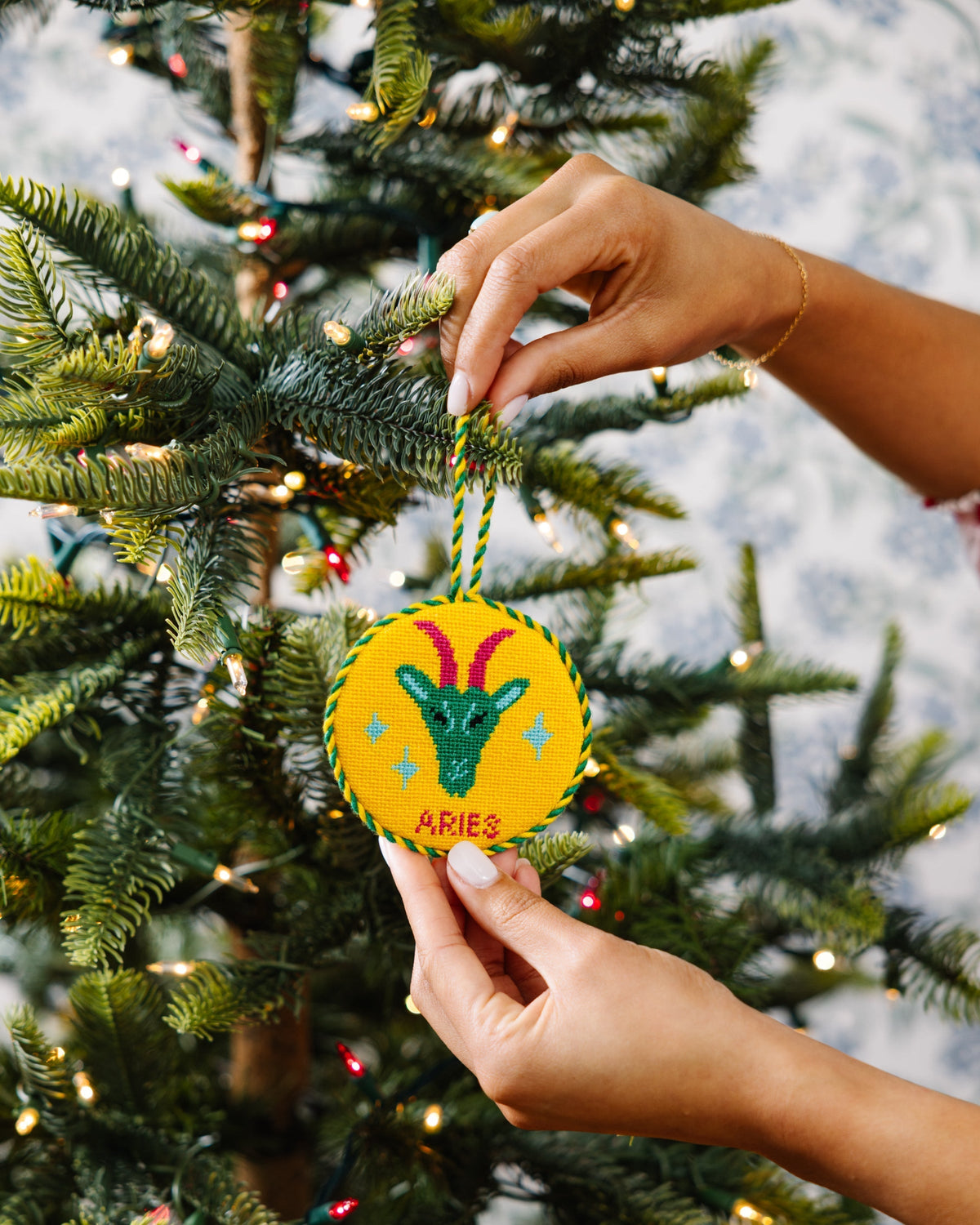 Hand holding a 'Aries' themed Christmas ornament against a decorated tree background