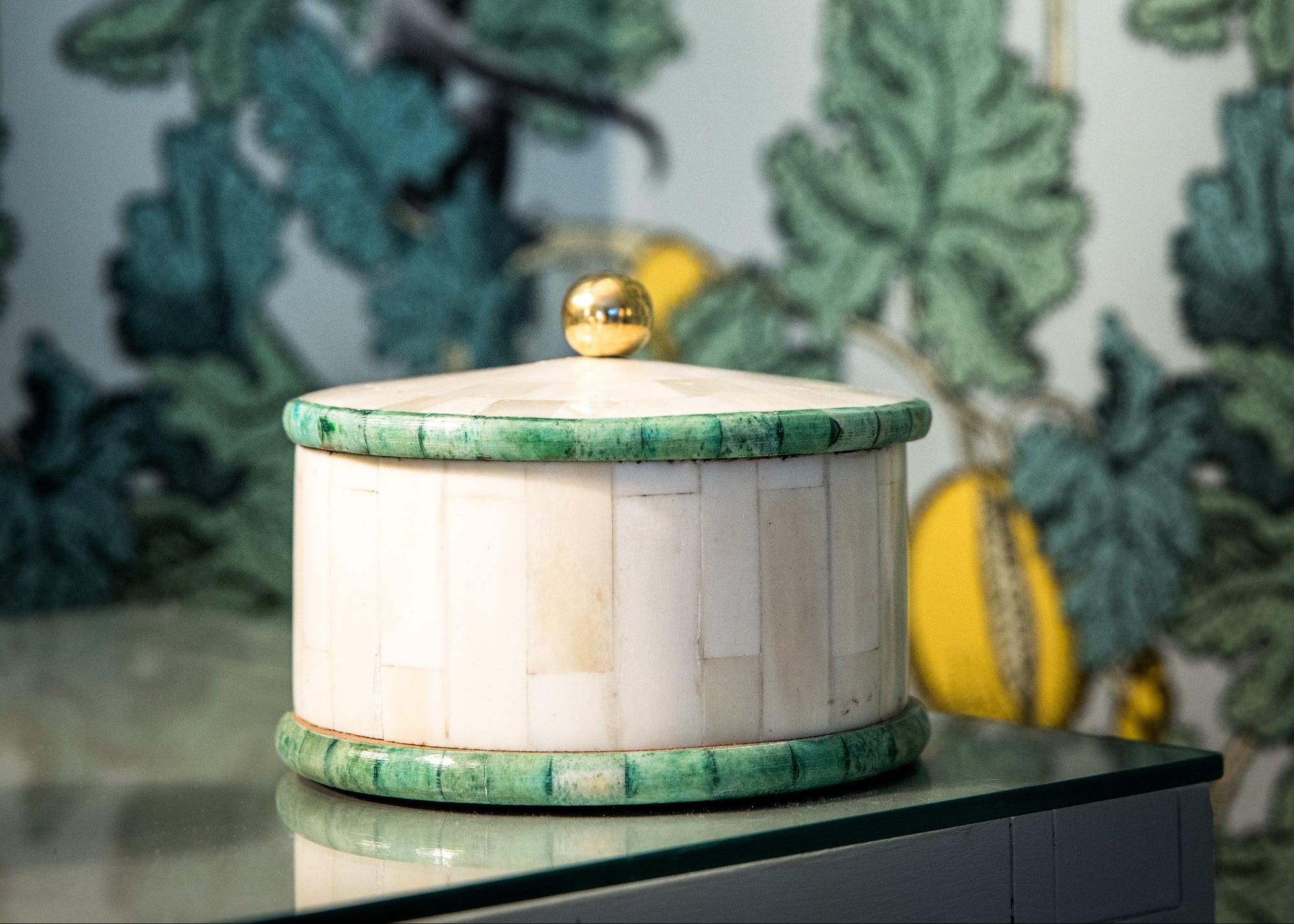 Decorative container with green lid on a surface against a leafy wallpaper background