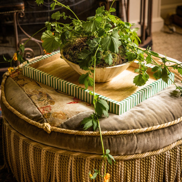 Round tassel ottoman with a plant on top of a tray in a cozy room setting with a fireplace in the background