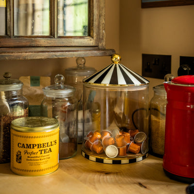 Glass jars filled with loose leaf tea, sugar and coffee pods on a kitchen counter next to a kettle. One jar is extra large with a circus top lid and matching base.