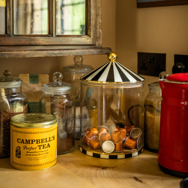 Glass jars filled with loose leaf tea, sugar and coffee pods on a kitchen counter next to a kettle. One jar is extra large with a circus top lid and matching base.
