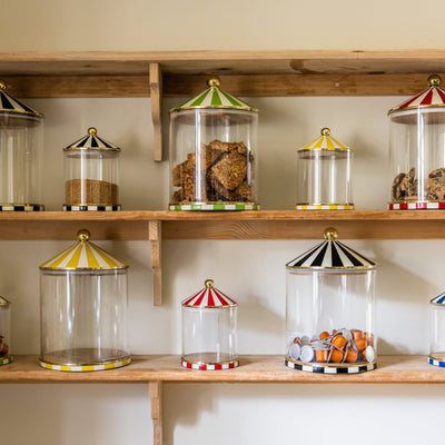 Wooden shelves with glass storage jars in two sizes featuring colourful circus lids on a neutral background.