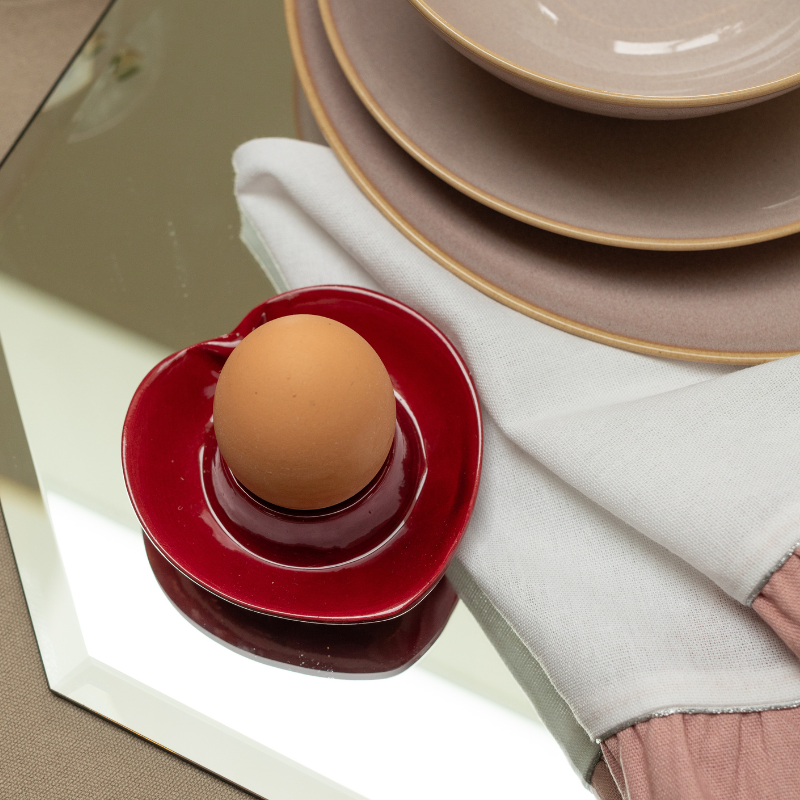 Brown egg on a red heart ceramic holder with beige plates and a white napkin in the background.