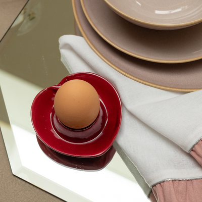 Brown egg on a red heart ceramic holder with beige plates and a white napkin in the background.