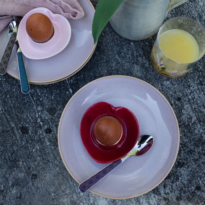 Eggs in red and pink cups holders on plates with plaid teaspoons on a stone table with a glass of juice and pitcher seen from above.