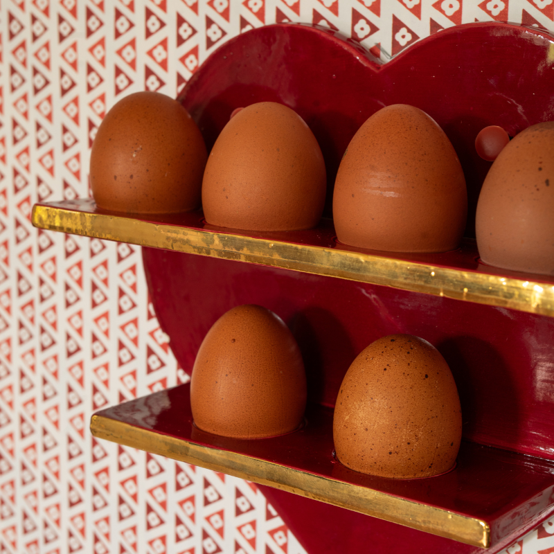 Red egg wall storage with two gold metallic shelves holding brown eggs against a patterned wallpaper background.