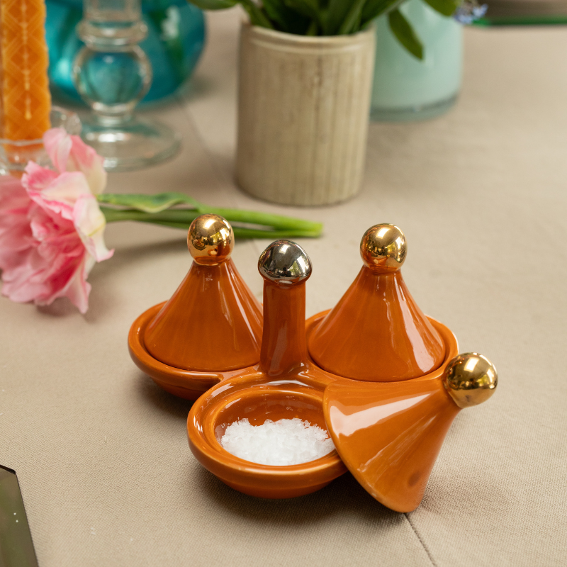 Set of three orange ceramic containers with gold knob lids on a tablecloth, with flowers and a vase in the background. One lid is balanced to reveal sea salt flakes inside.
