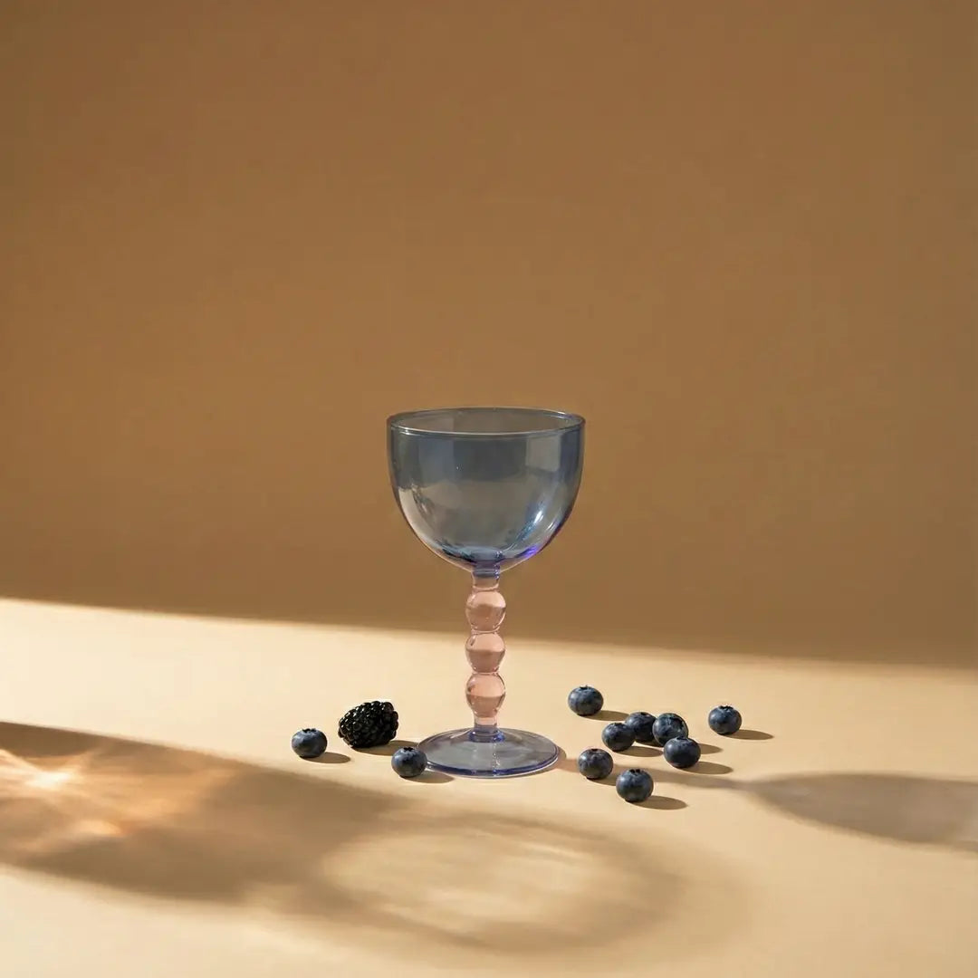 Blue glass goblet with a pink stem on a beige surface with scattered berries