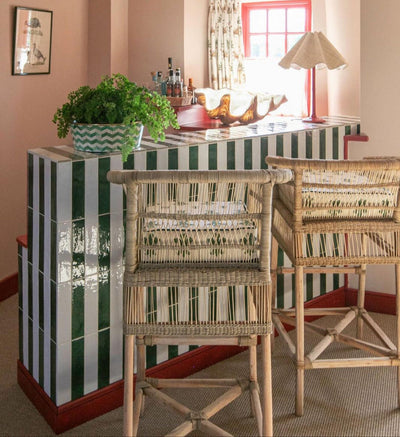 A green and white metro tile home bar with zigzag planter and fern on top along with decorative clam, drink bottles, ruffle lampshade and rattan bar stool chairs.