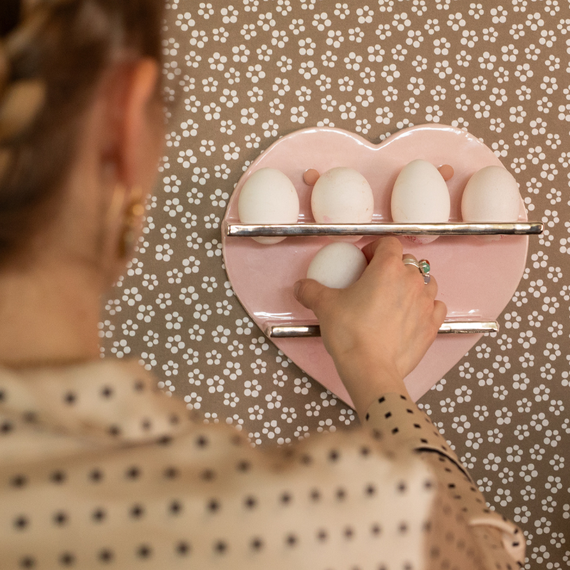 Person placing a white egg on the bottom shelf of a pink heart-shaped egg holder with eggs already on the top shelf against a beige floral-patterned wall.