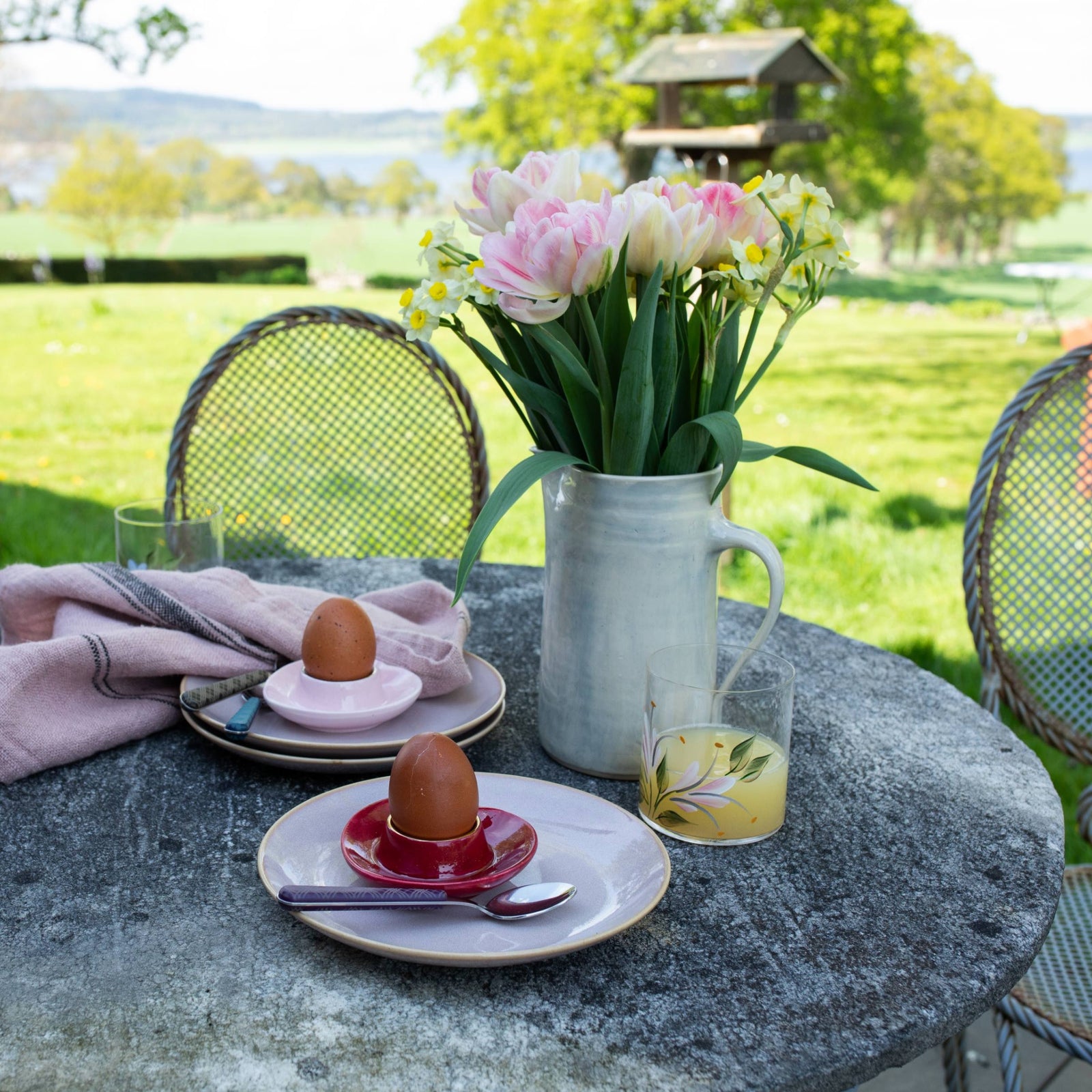 Outdoor setting with a table set for breakfast, featuring eggs in egg cups on plates with plaid teaspoons, a glass of orange juice, napkins and a vase of tulip flowers.