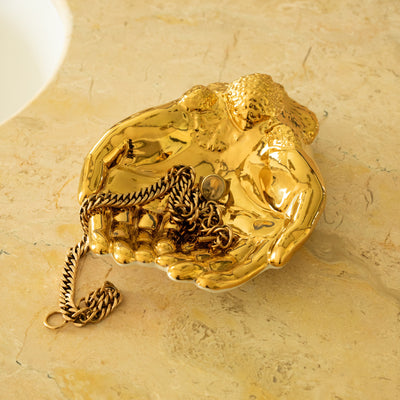 Gold decorative hand shaped dish holding jewellery on a beige marble surface
