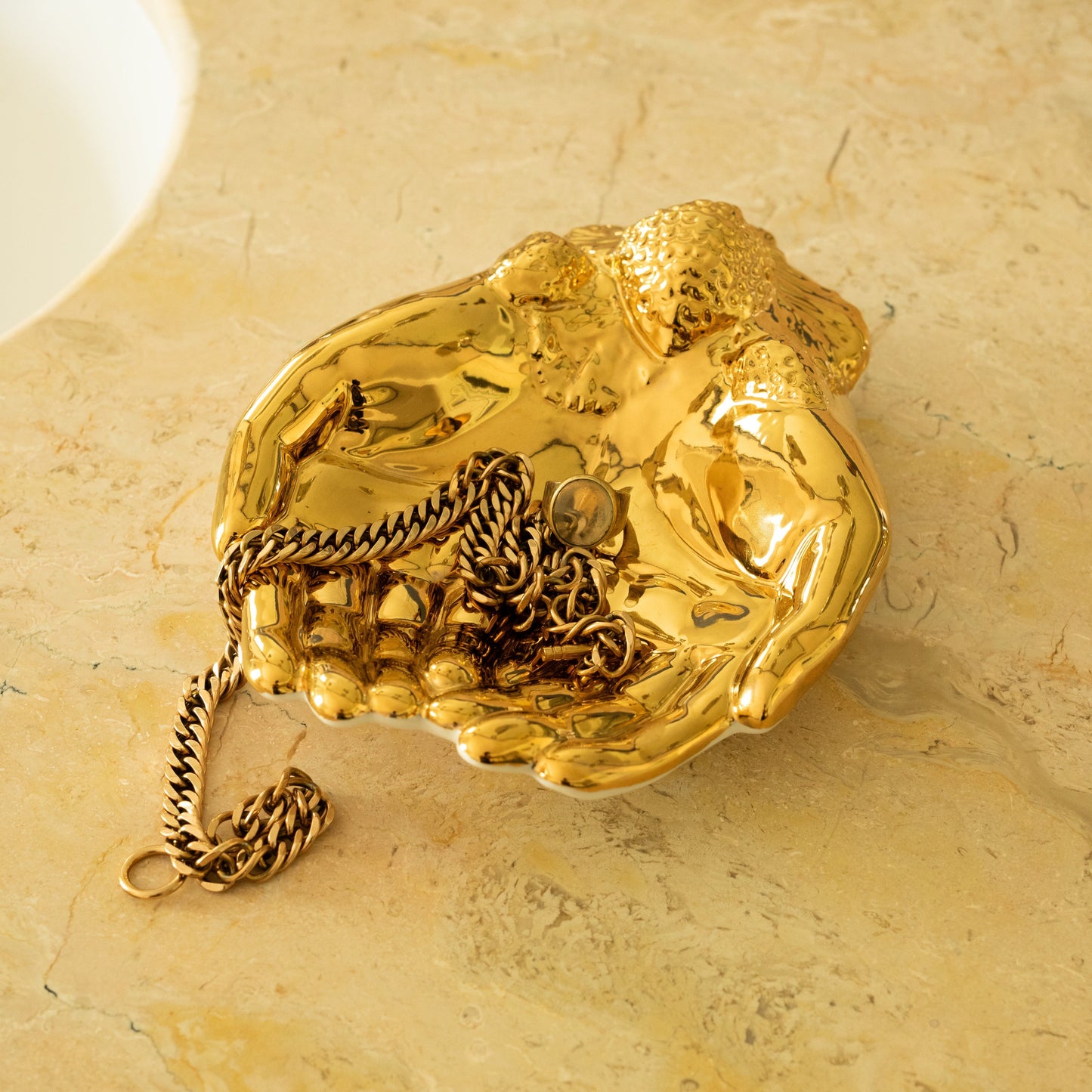 Gold decorative hand shaped dish holding jewellery on a beige marble surface