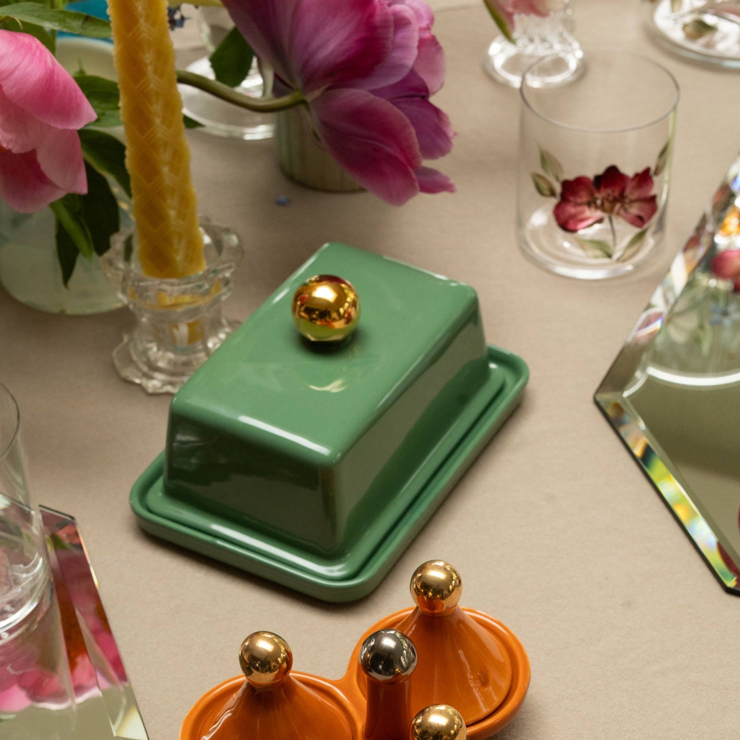 Decorative items on a table including a teal green butter tray with gold ball lid, orange condiment caddy with gold balls on its lids, a yellow tartan embossed taper candle, hand painted floral glass tumblers, and pink flowers.