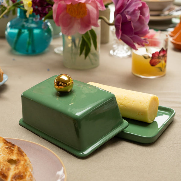 Green butter dish open to reveal butter inside with a gold knob on the lid amongst flowers and a hand painted floral glass in the background on a tablescape.