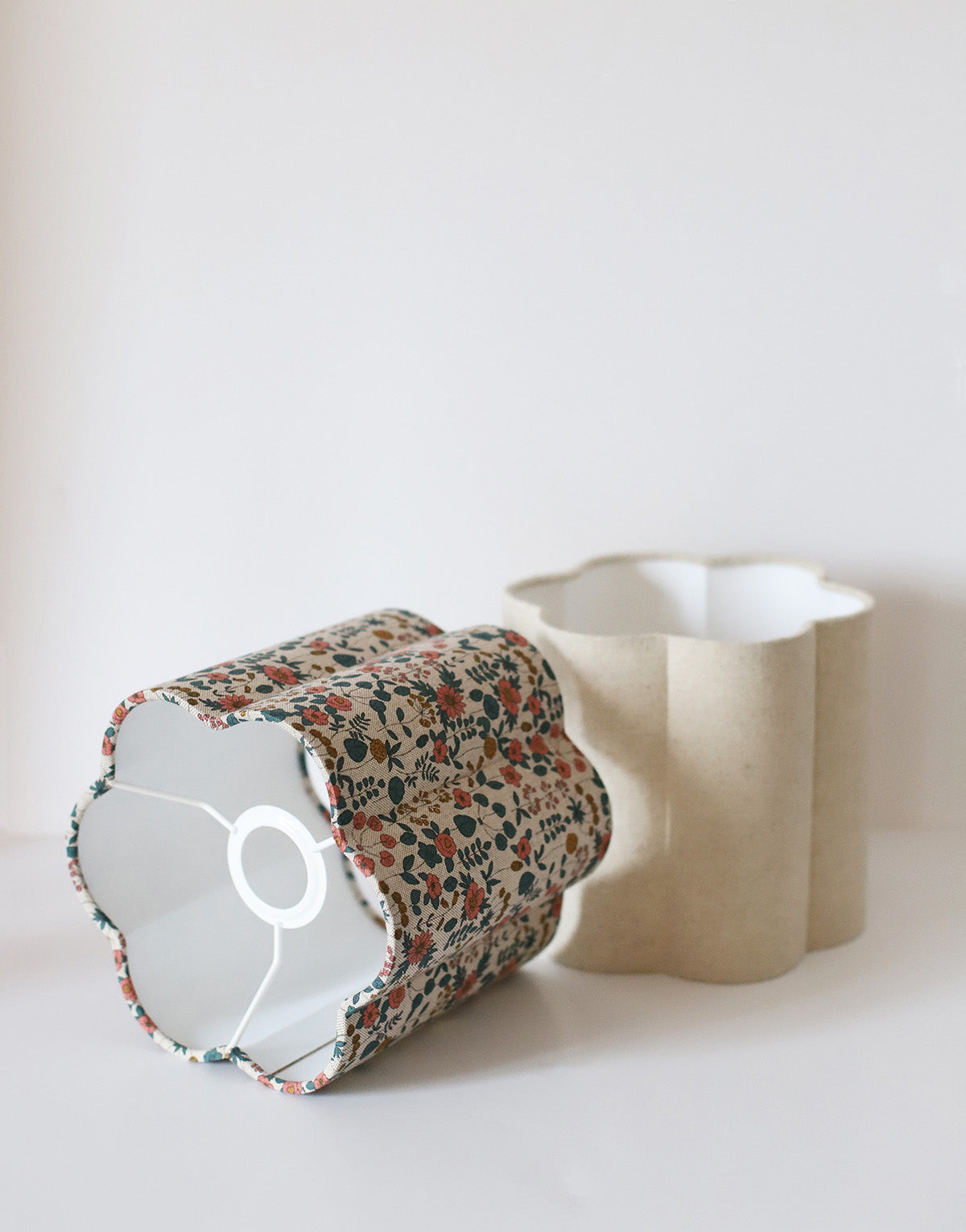 Abat-jour fleur Flora en coton beige fleuri, décor floral sur fond clair, fait main, posé à côté d’un support blanc.