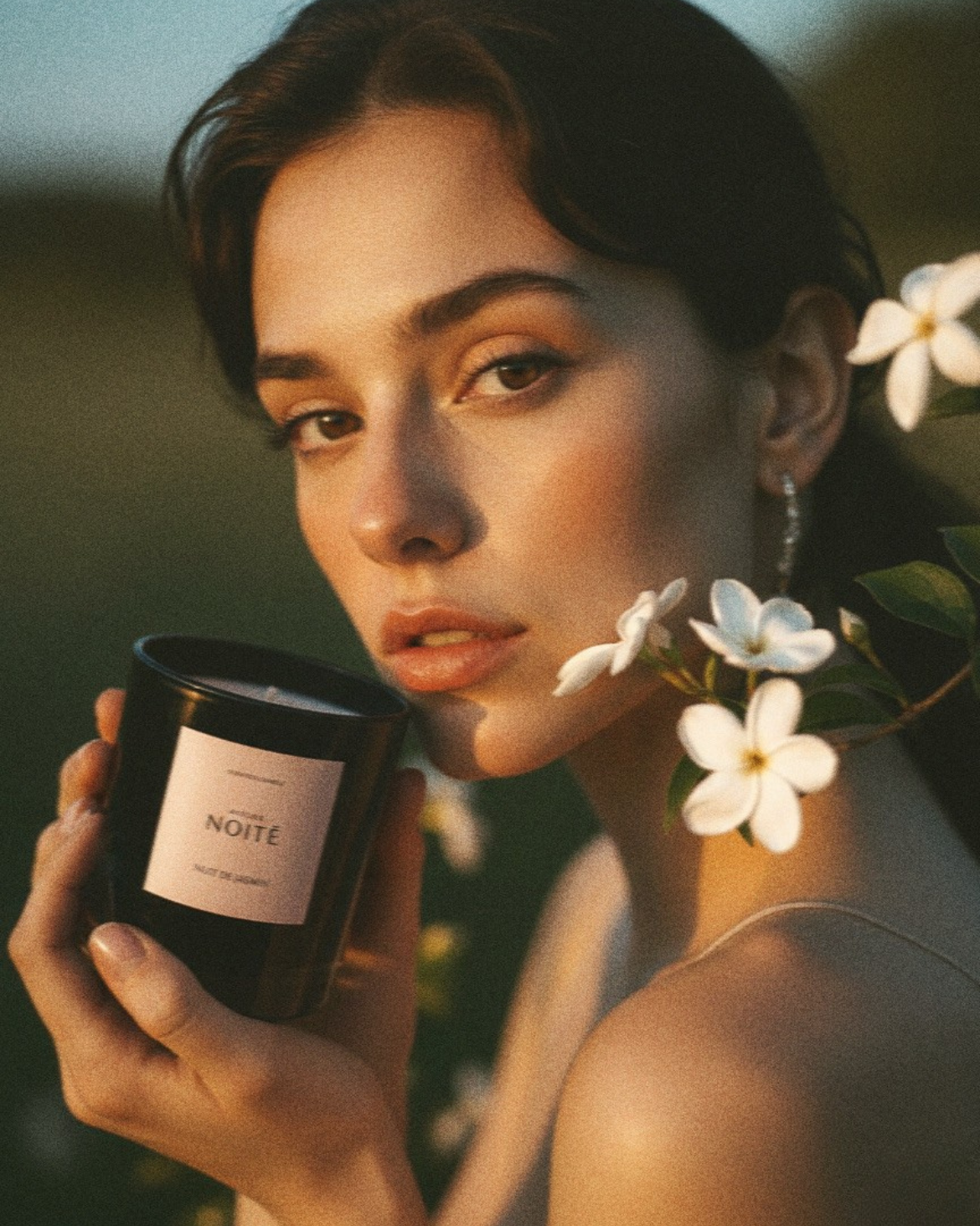 Woman holding a candle labeled 'Atelier Noite' with flowers around her