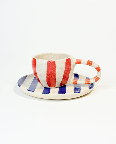 A hand-painted ceramic mug with vertical red and white stripes, accompanied by a round plate with horizontal blue and white stripes.