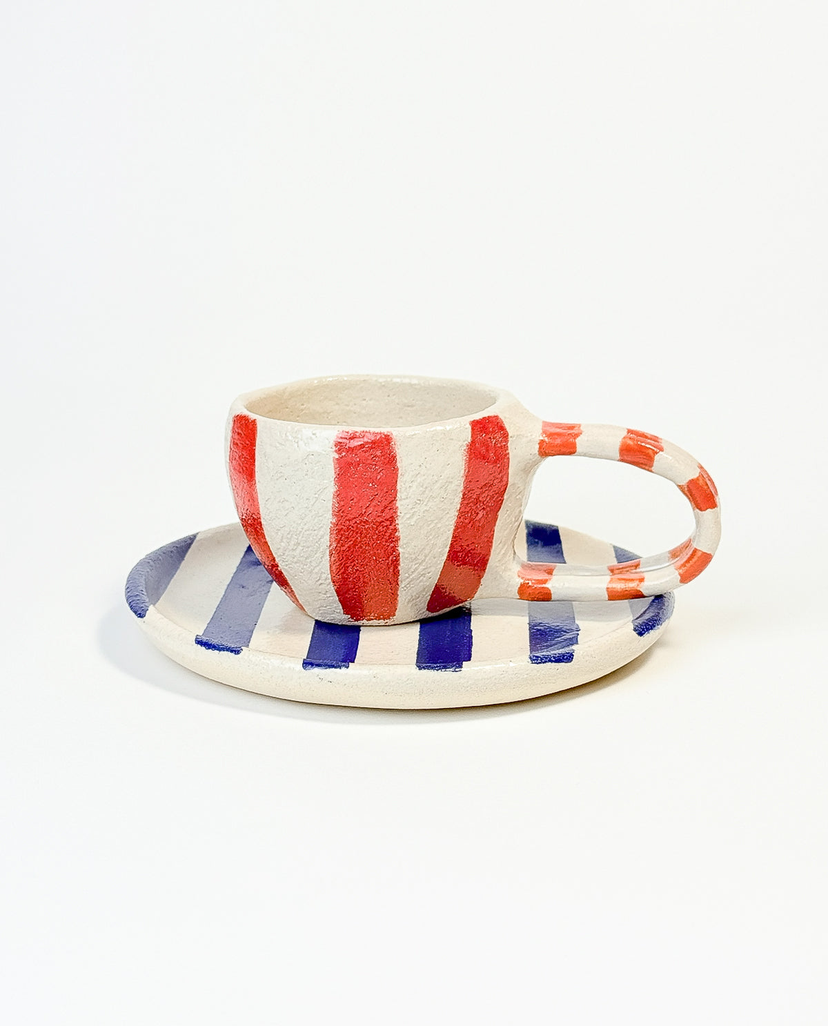 A hand-painted ceramic mug with vertical red and white stripes, accompanied by a round plate with horizontal blue and white stripes.