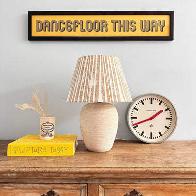 Bold stripe lampshade in warm neutral tones displayed on a rustic wooden sideboard, styled with a clock and books. A modern empire lampshade that combines striped lighting design with timeless elegance.