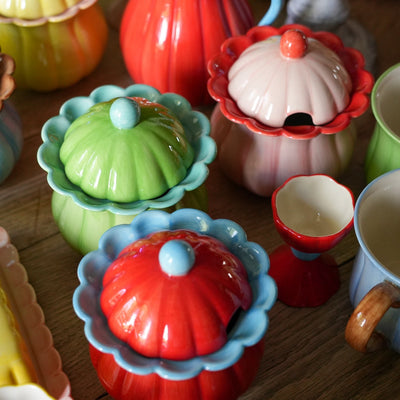 A collection of colorful, pumpkin-shaped ceramic containers and serving dishes arranged on a wooden table. The set features vibrant red, green, blue, yellow, and pink items with scalloped edges and glossy finishes.