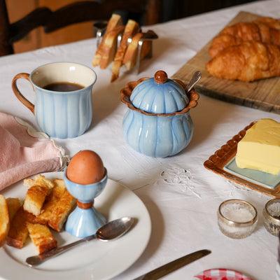 A breakfast table setting with a blue ribbed mug of coffee, a blue lidded sugar bowl, croissants on a wooden board, butter on a plate, toast on a white plate with an egg in an egg cup, and a pink cloth napkin on a white tablecloth.