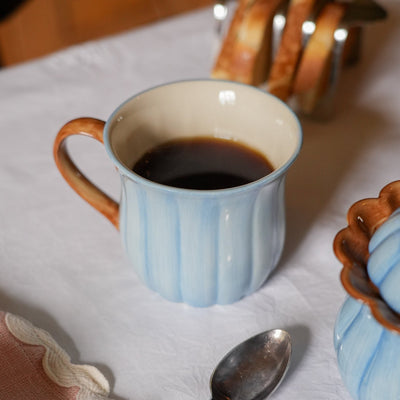 A light blue and white ribbed ceramic mug filled with black coffee, set on a white tablecloth with a spoon in the foreground and bread in the background.