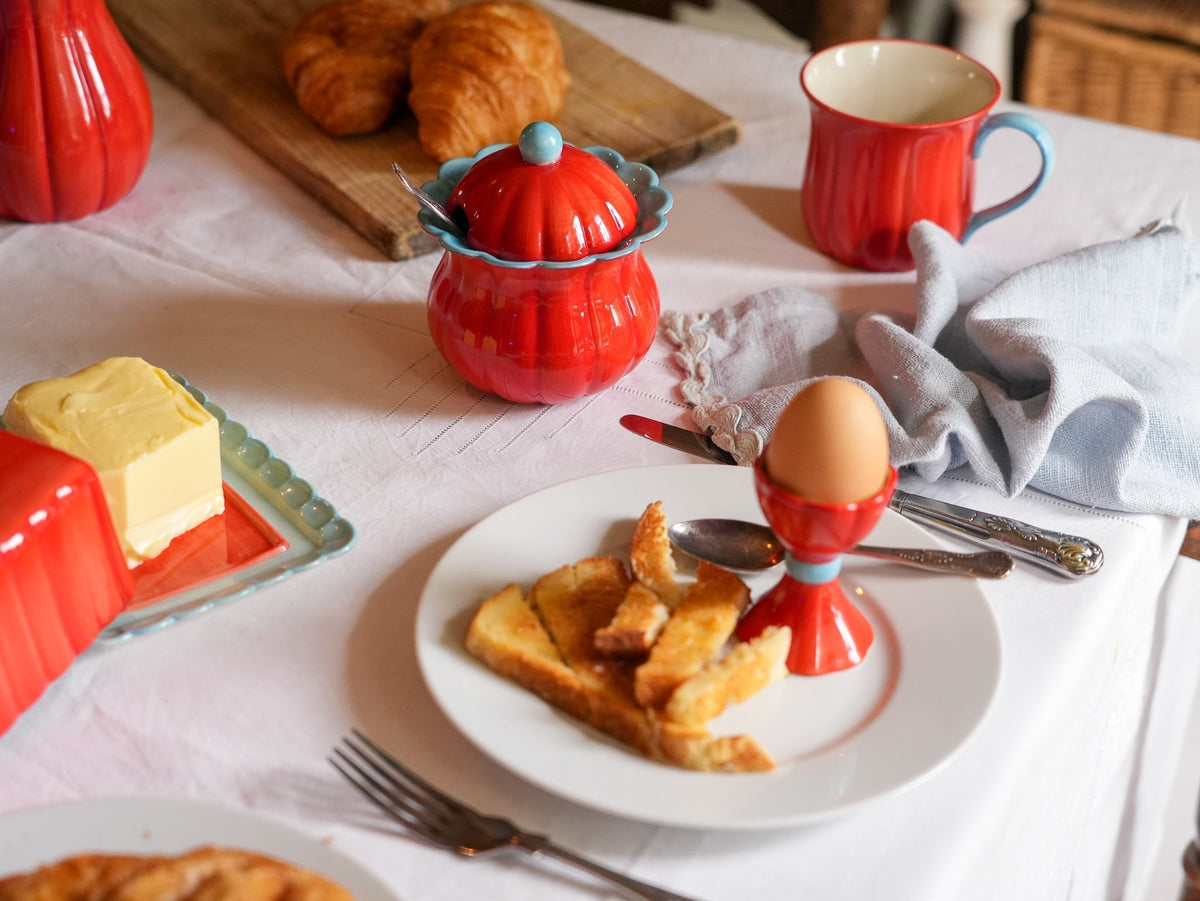 A styled breakfast table scene with red ceramic dishes and mugs, a plate of toast and an egg in an egg cup, butter, croissants, and a white tablecloth.