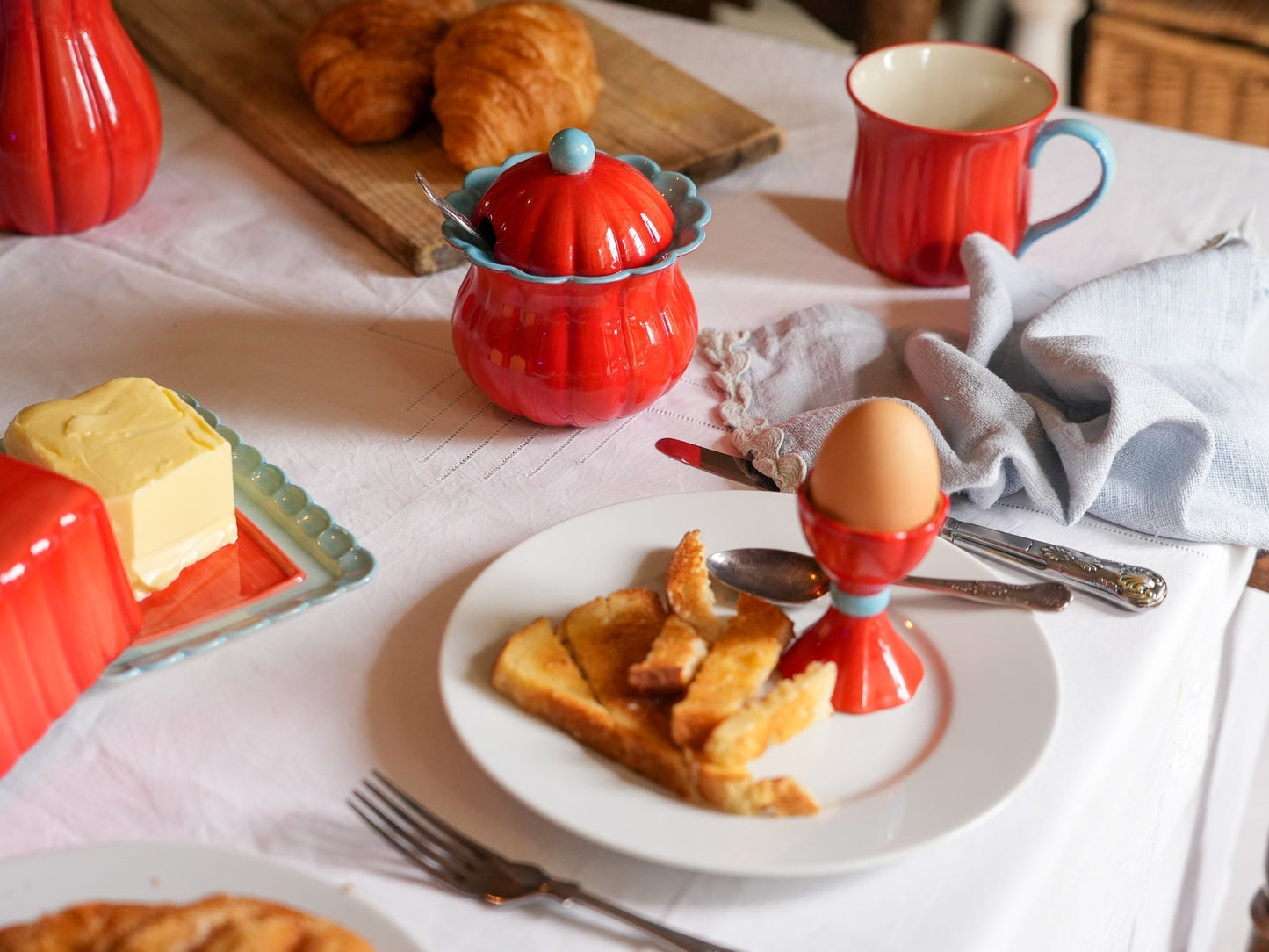 A styled breakfast table scene with red ceramic dishes and mugs, a plate of toast and an egg in an egg cup, butter, croissants, and a white tablecloth.